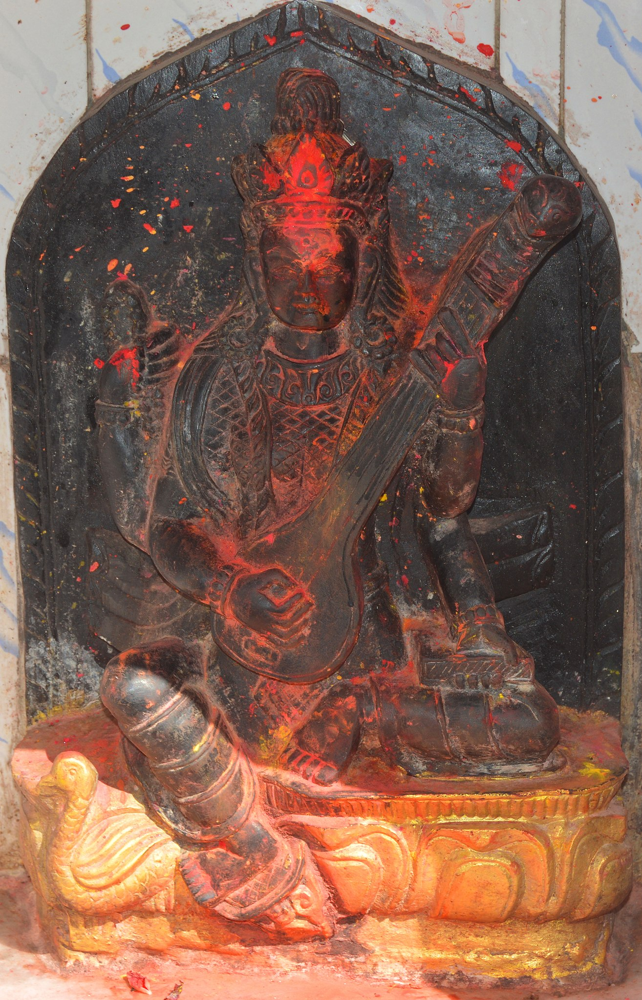 A carved stone statue of Saraswati, painted with red vermilion, seated with her veena at a Kathmandu temple; symbolizes her role as goddess of learning and creative inspiration.