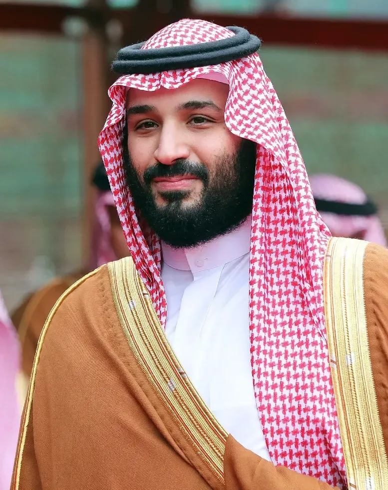 Mohammed bin Salman in traditional Saudi dress with red-and-white headscarf and gold-trimmed bisht, smiling