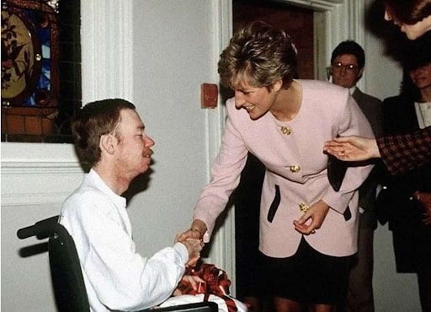 Princess Diana warmly shakes hands with an AIDS patient, wearing a light pink blazer and smiling with compassion, surrounded by onlookers in a clinical setting.
