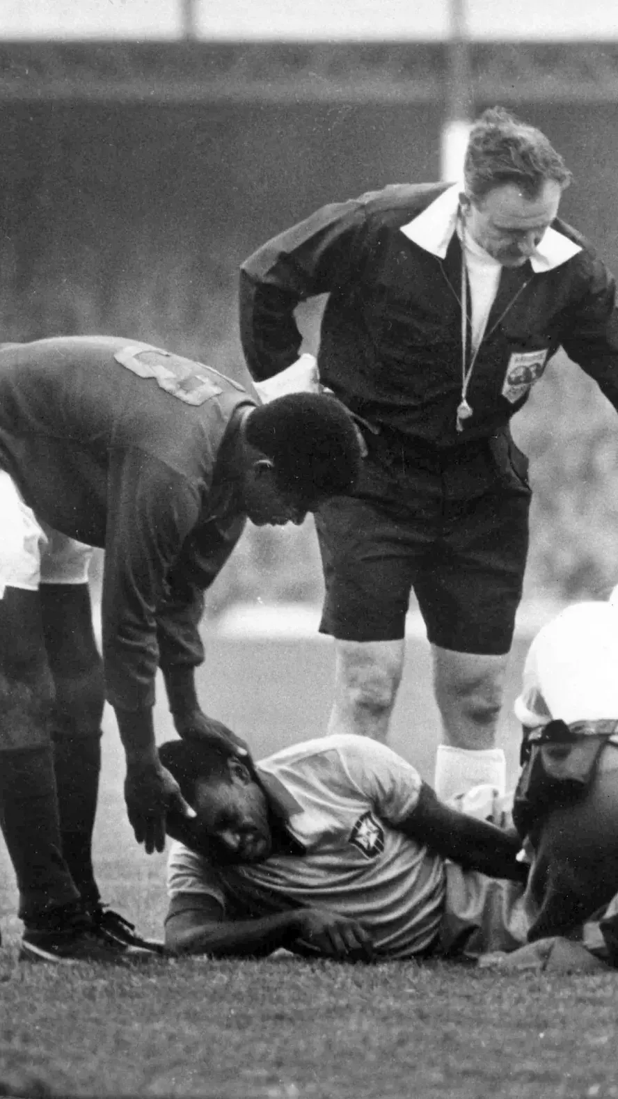 Pelé lying injured on the pitch during the 1966 World Cup as officials and teammates attend to him, symbolizing the cost of joy and creative play under pressure.