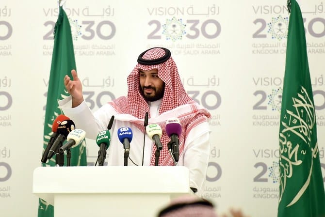 Mohammed bin Salman Al Saud speaking at a podium with the Vision 2030 logo and Saudi flag in the background.