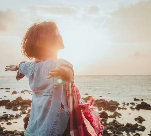 Woman facing the ocean with arms wide open, embodying the feeling of the wish fulfilled under golden sunlight — Law of Assumption visualization.