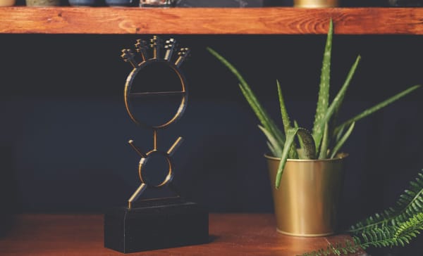 Bob Proctor Stick Man metal sculpture on a wooden shelf beside a potted aloe plant.