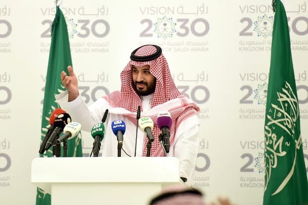 Mohammed bin Salman Al Saud speaking at a podium with the Vision 2030 logo and Saudi flag in the background.